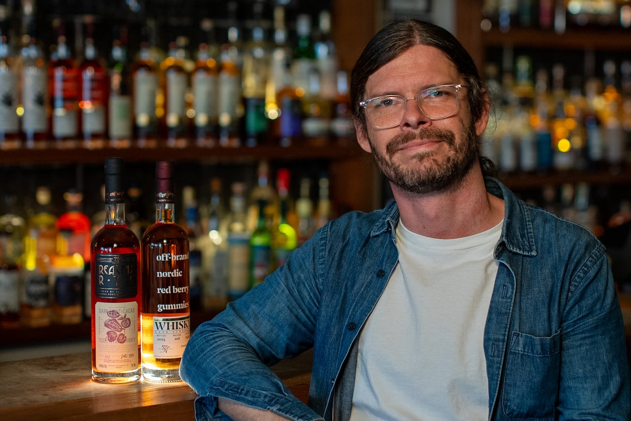 a man with glasses and a demin shirt over a white tshirt smiles at the camera. he's sitting at a bar rail beside two bottles of whiskey