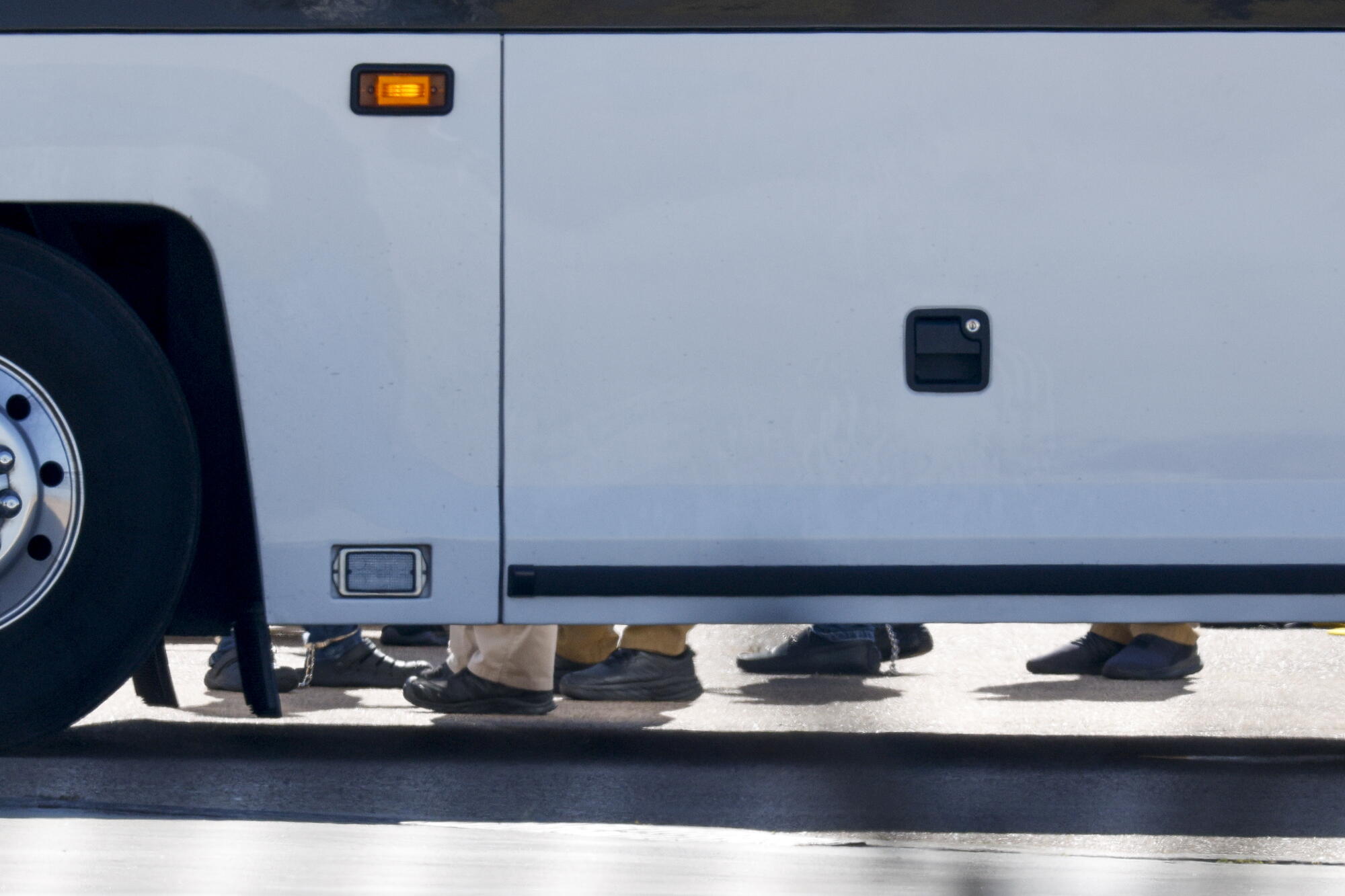 The feet of restrained people standing on the other side of a white vehicle 