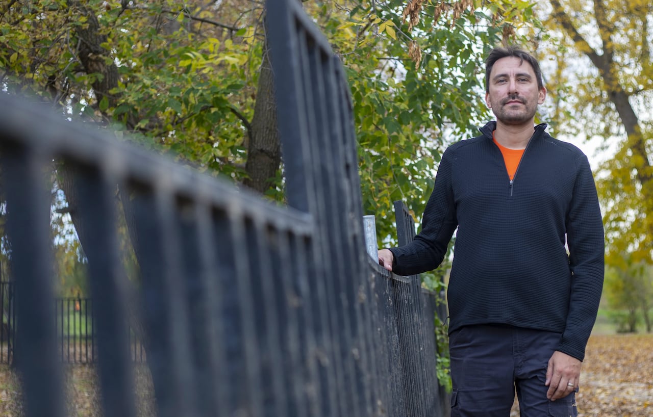 A man stands beside a black metal gate in a treed area.