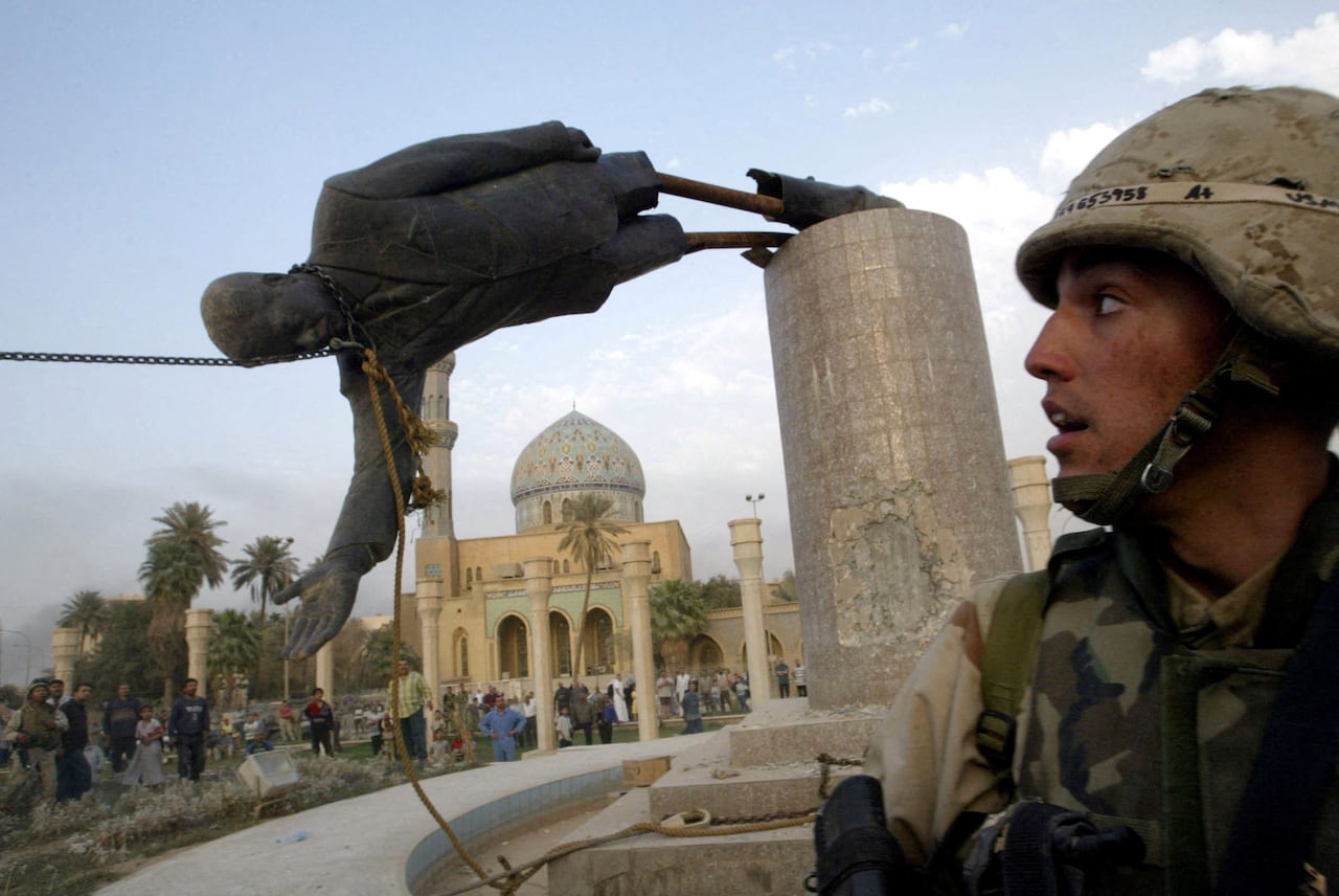 A helmeted soldier in uniform looks up at a statue being toppled.