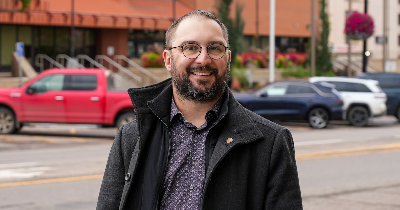 A politician smiles for a photo wearing a black jacket. There are vehicles in the background parked in front of a brick building.