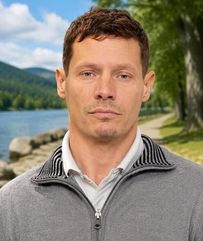 A man with brown hair in a grey sweater stands in front of a tree and path.