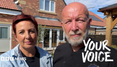 Elissa and Johnnie stand in front of a modern brick house with red‑tiled roofing and solar panels. Elissa wears a light blue shirt, the Johnnie a black shirt with safety glasses tucked into the pocket. The house has white‑framed windows, a light‑coloured door, and a clear blue sky overhead.