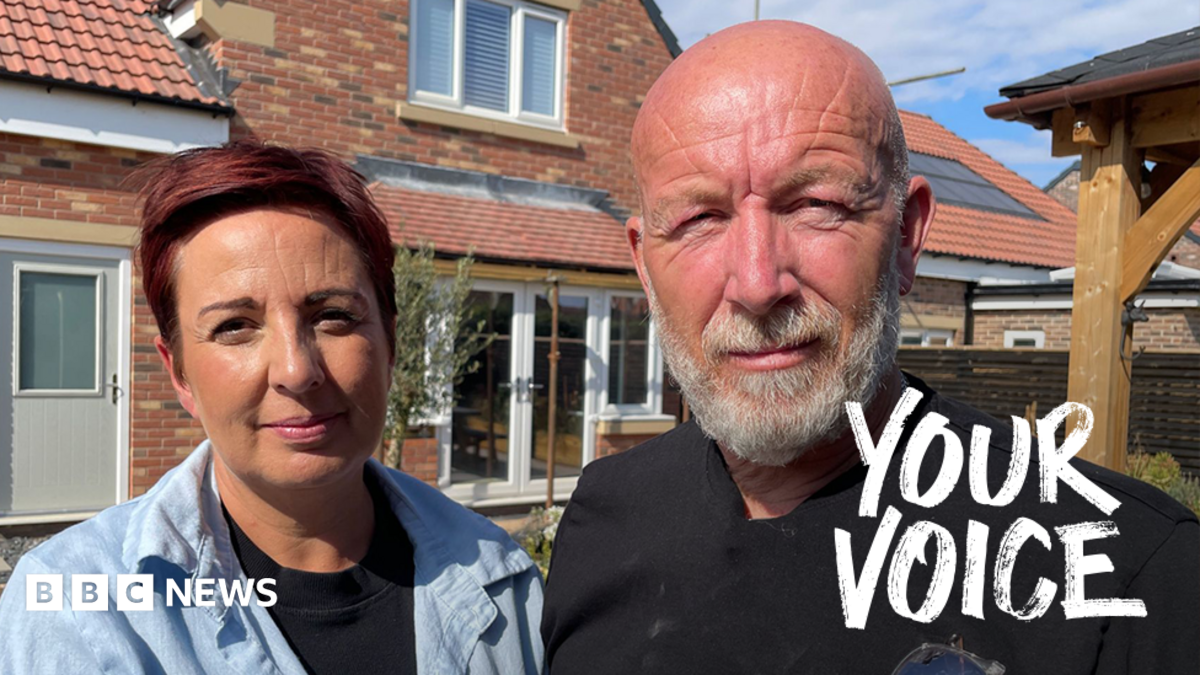 Elissa and Johnnie stand in front of a modern brick house with red‑tiled roofing and solar panels. Elissa wears a light blue shirt, the Johnnie a black shirt with safety glasses tucked into the pocket. The house has white‑framed windows, a light‑coloured door, and a clear blue sky overhead.