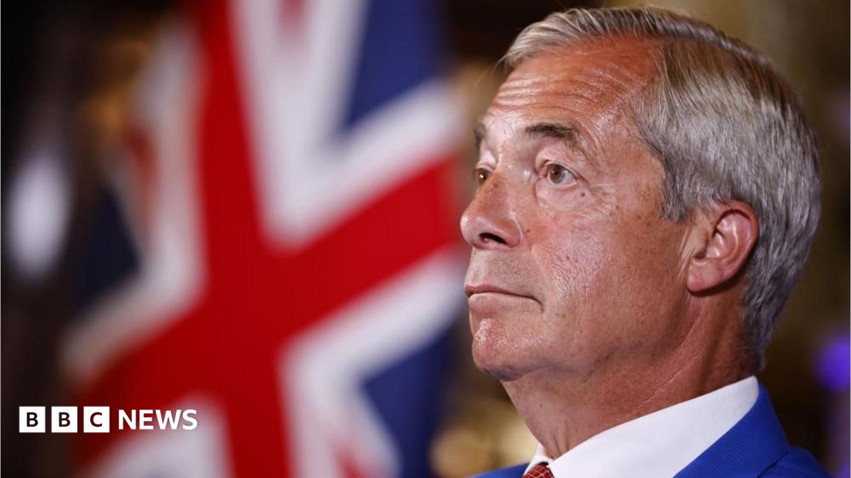 Nigel Farage is wearing a blue suit, white shirt and a tie. He has short grey hair. A Union flag can be seen in the background.