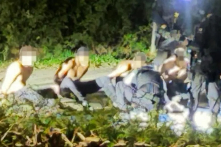 Five men with their faces blurred sitting bow-legged on the ground while handcuffed next to two policemen in black clothes.