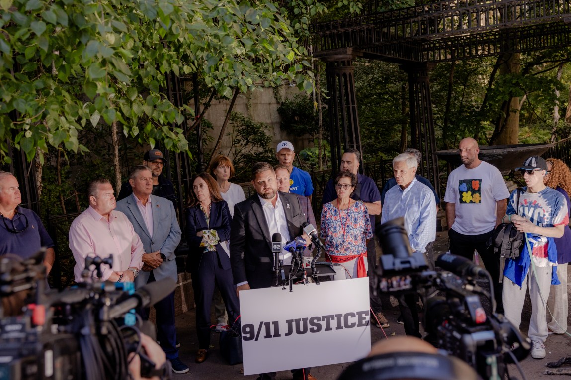 A man stands at a podium speaking into TV station microphones. A crowd of people stands with him, and in front of them is a sign that reads “9/11 Justice.”