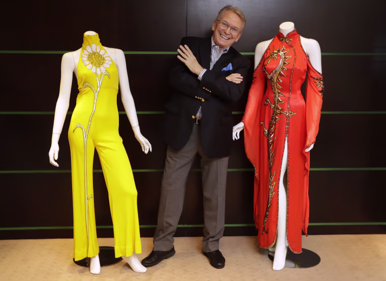 Bob Mackie poses with two of his iconic designs, a scarlet red satin gown worn by Cher in 1975, right, and a marigold jersey jumpsuit worn by Cher between 1971-1976, left, in London, in 2018.