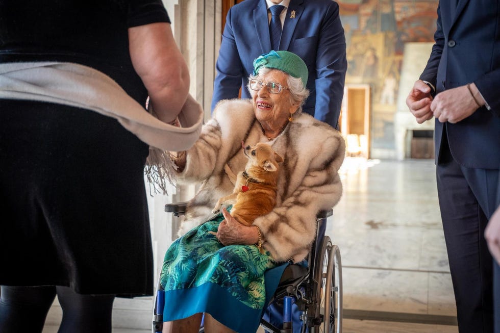 Elderly woman in a wheelchair holding a small dog, interacting with other individuals.