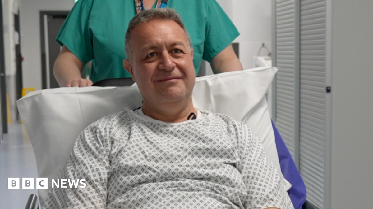 Steve wears a hospital gown and sits up on a gurney. He is propped up on a pillow and is wearing a patterned hospital gown in black and white. He is being pushed on his way to have his operation