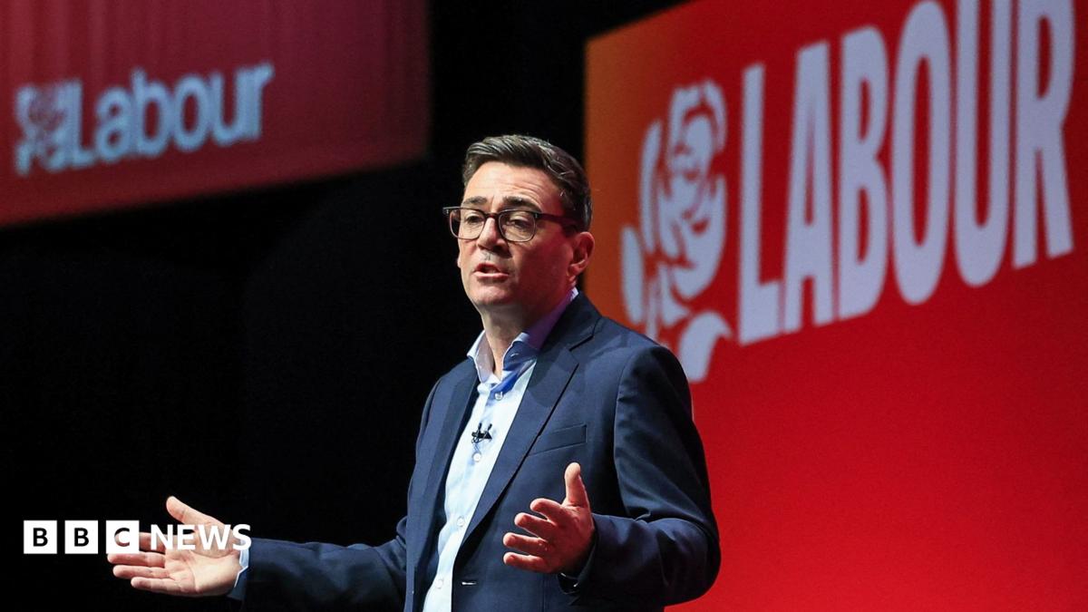 Andy Burnham is wearing a dark-coloured jacket, a white shirt and glasses. He is stood in front of a red backdrop.