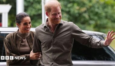 Meghan holding onto Harry's arm, while Harry waves his hand