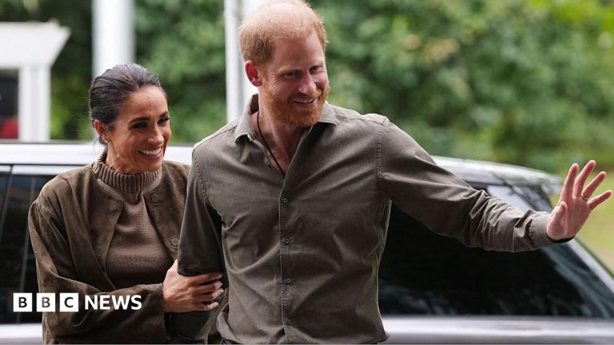Meghan holding onto Harry's arm, while Harry waves his hand