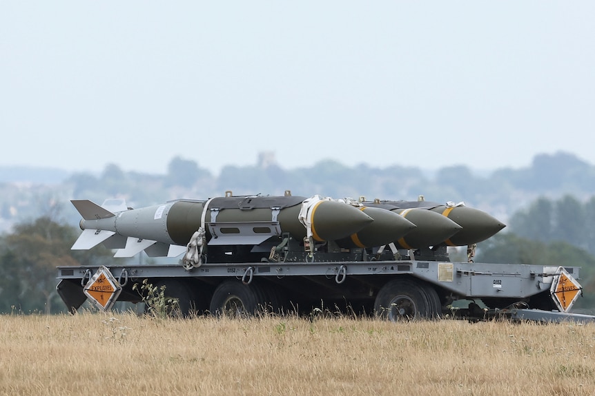 Three large, olive green rocket munitions sit on the back of a grey trailer trolley in an open field.