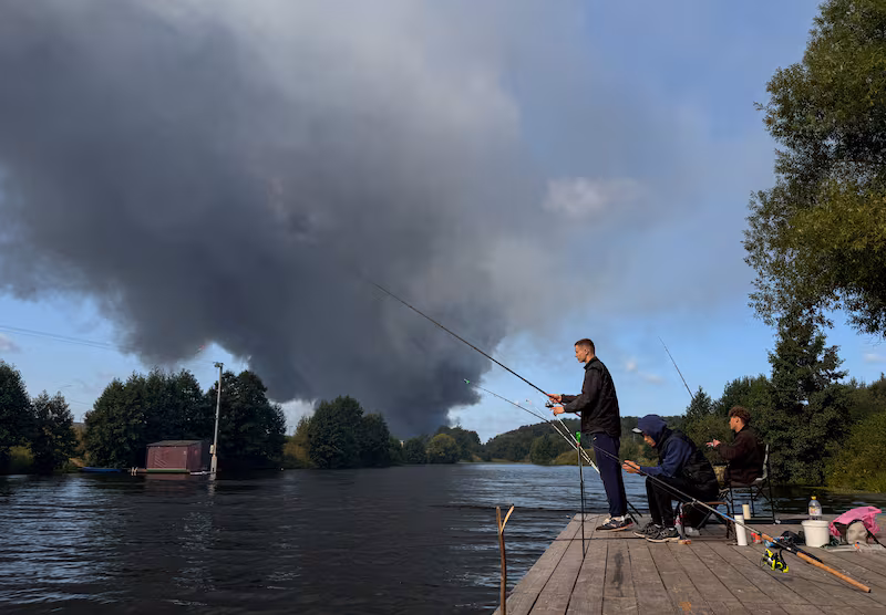 People fish from a pier as smoke billows from the burning Wildberries warehouses following a Ukrainian drone attack, according to the regional governor, in the course of the Russia-Ukraine conflict, in Koledino, Moscow Region, Russia August 16, 2026. Picture taken with mobile phone. REUTERS/Stringer     TPX IMAGES OF THE DAY