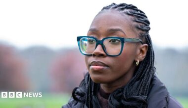 Close-up portrait of Christine outdoors wearing large blue-framed glasses, small hoop earrings and a dark padded coat. She is looking slightly away from the camera, with braided hair visible. The background is softly blurred, showing a green field and trees in muted colours.