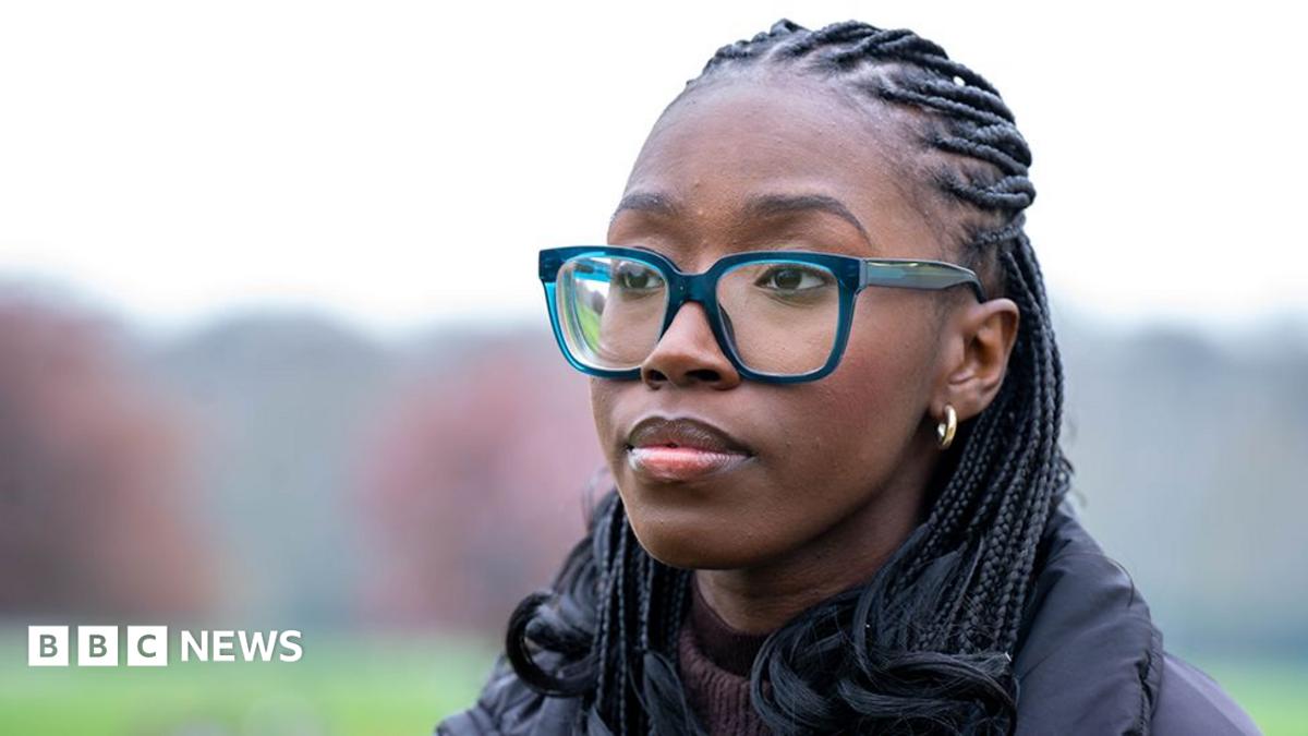 Close-up portrait of Christine outdoors wearing large blue-framed glasses, small hoop earrings and a dark padded coat. She is looking slightly away from the camera, with braided hair visible. The background is softly blurred, showing a green field and trees in muted colours.