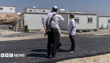 Three Israeli settlers stand outside houses after the reopening of a settlement at Ganim in the Israeli-occupied West Bank. One is a man armed with a rifle. Photo: August 2026