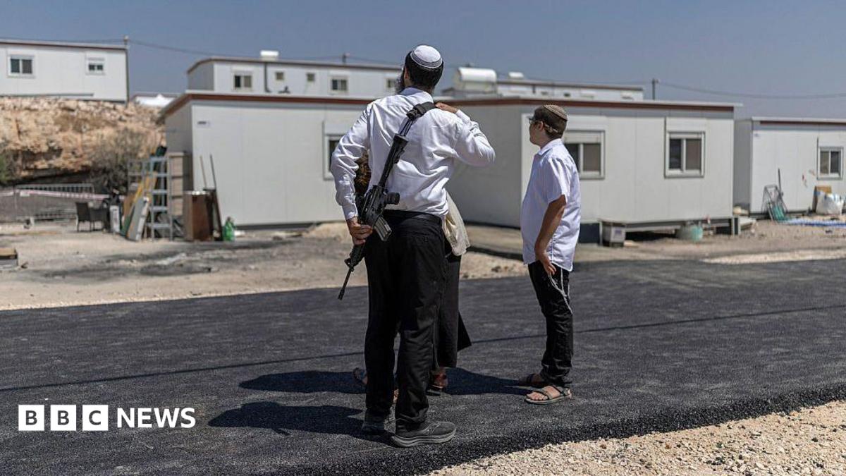 Three Israeli settlers stand outside houses after the reopening of a settlement at Ganim in the Israeli-occupied West Bank. One is a man armed with a rifle. Photo: August 2026