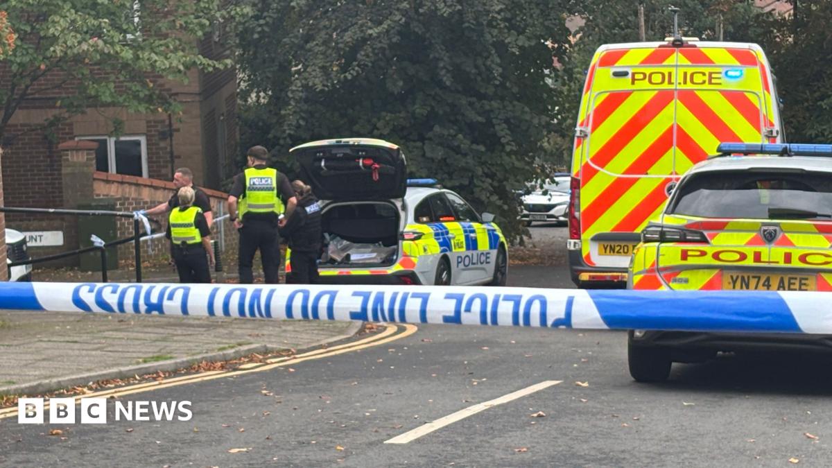 Four police officers are visible in the distance. They are standing at the back of a police car, which, along with another police car and van, are behind a police cordon