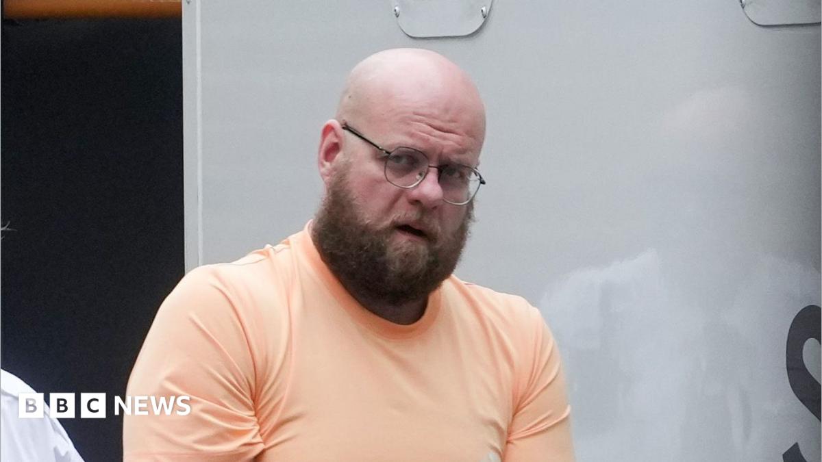 A man wearing an orange t-shirt. He is being led from a prison van by a custody officer.