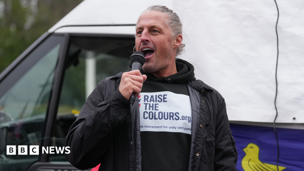 Image shows a man with tied back blonde/grey hair holding a black microphone.  He is talking and smiling, wearing a black jacket and a black hoodie with a white logo that says in blue 'Raise The Colours'