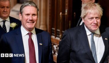 Keir Starmer on the left, and Boris Johnson on the right, walking together in the Houses of Parliament in 2024. They are smiling and both wear white shirts, a lightly patterned tie, and blue suit jackets.