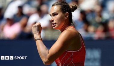 Aryna Sabalenka, wearing an orange sleeveless dress, stands side on and raises a clenched left fist