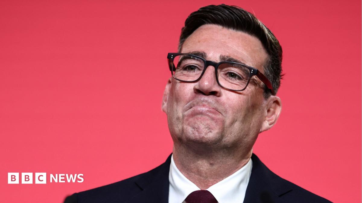 Andy Burnham looking pleased as he reacts to becoming Labour leader. He is wearing black rimmed glasses and a dark suit and tie, against a red background.