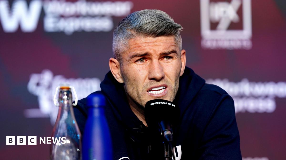 A man in his 30s, with grey hair and wearing a blue hoody, speaks into a black microphone at a press conference.