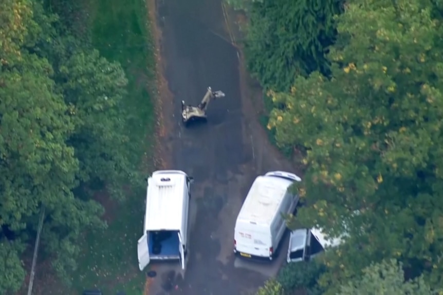 Two white vans, with one of its rear doors open, seen on a roadway in between green trees and a tan bomb disposal robot.