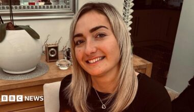 Maddy Cusack pictured smiling at home. She has long blonde hair worn loose and wears a silver heart necklace over a black top. She's pictured inside, in front of a side board with ornaments and plants