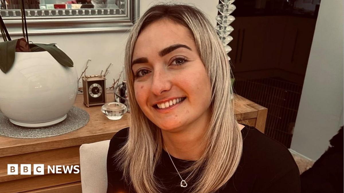 Maddy Cusack pictured smiling at home. She has long blonde hair worn loose and wears a silver heart necklace over a black top. She's pictured inside, in front of a side board with ornaments and plants