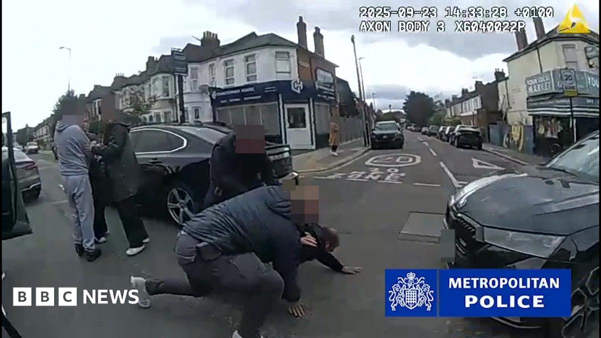 Police bodycam footage shows two officers arresting a man, who is on his hands and knees on the road, beside some parked cars in a residential street. To their left, are three other individuals, their faces blurred.