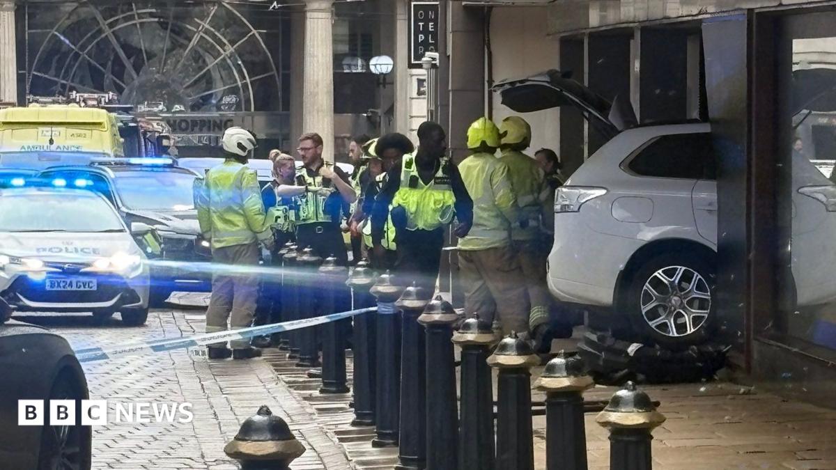 A white car can be seen halfway in a building's glass frontage with firefighters and emergency crews standing by it and debris can be seen on the floor. Blue flashing lights from police vehicles can also be seen.