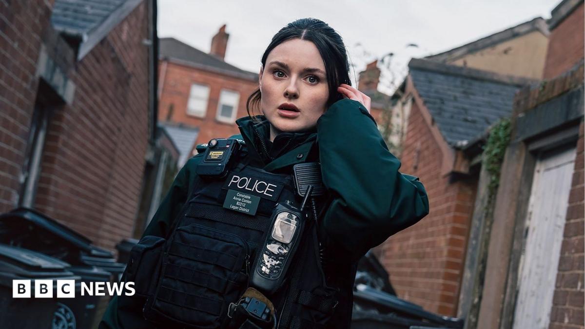 A young woman with brunette hair dressed in a dark green police uniform which includes a body-worn camera, stab vest and walkie-talkie. She's standing in an alleyway with red brick houses and bins alongside her. She's holding an ear piece to her left ear. She has a stunned impression on her face.