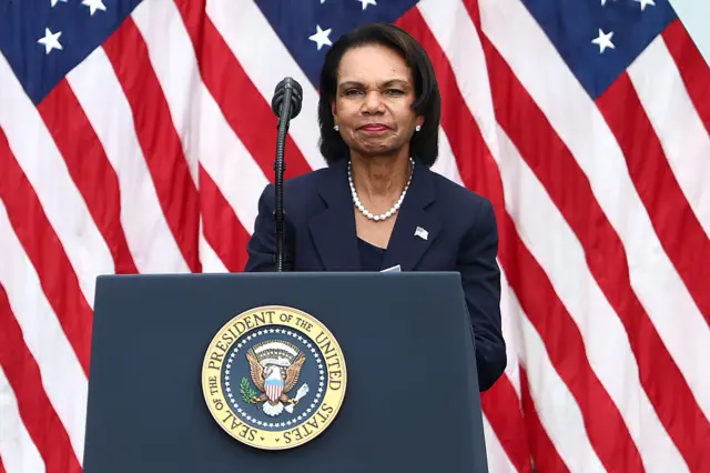 Former Secretary of State Condoleeza Rice delivers a speech in a black suit and shirt whilst standing behind a lectern bearing the sigil of the US president, several US flags behind her