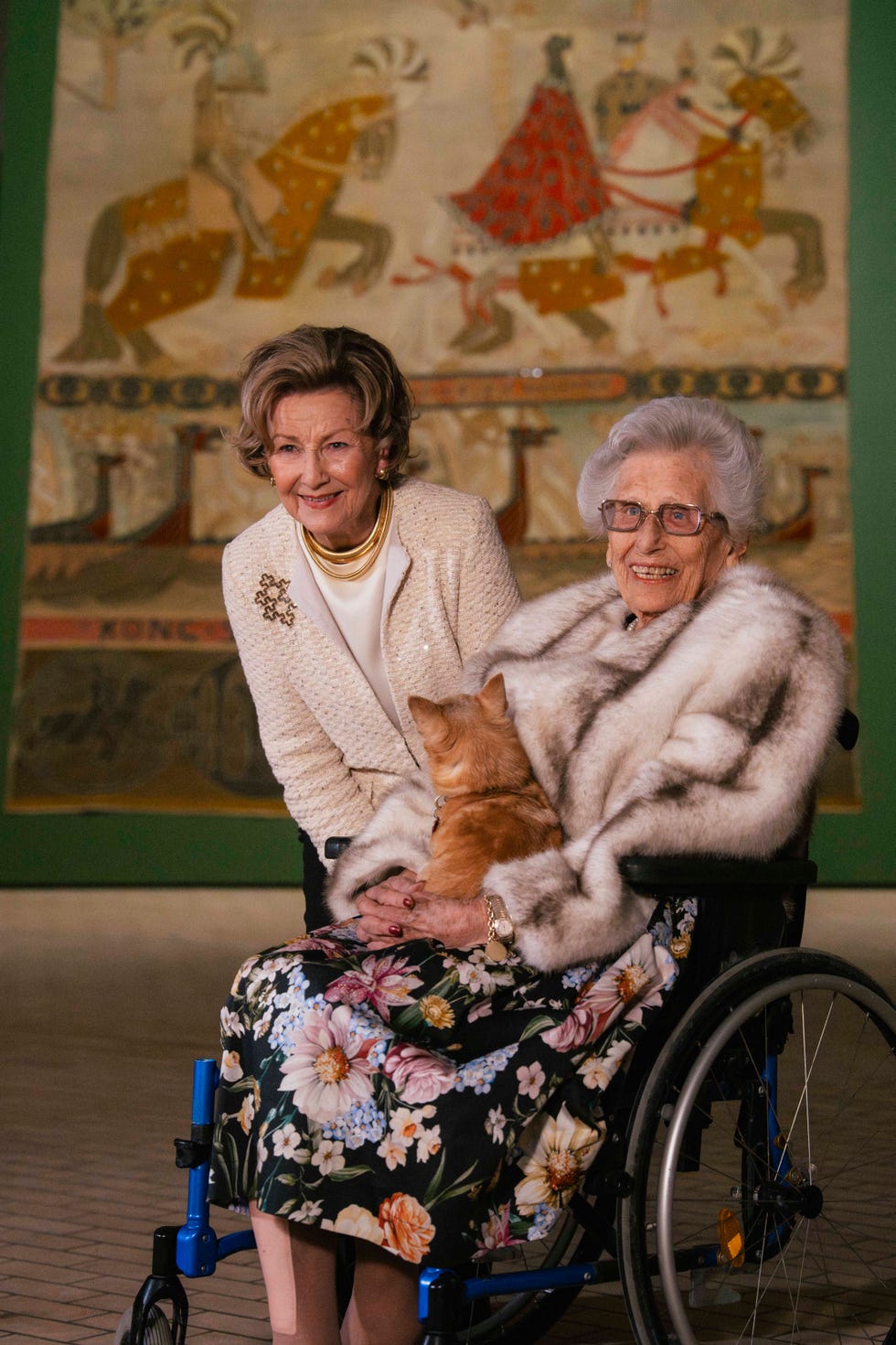 Two women, one seated in a wheelchair, with a cat in their lap, in front of a tapestry.