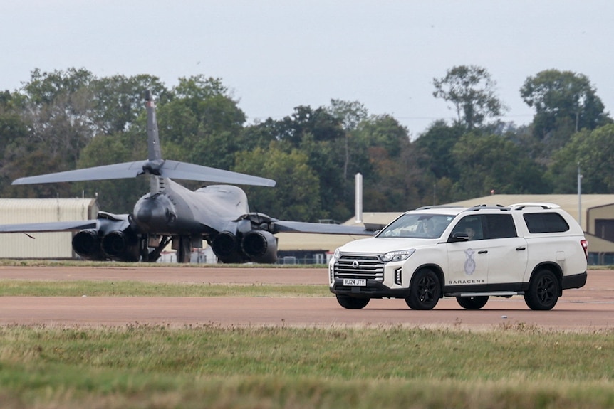 A large, dark grey military plane seen on a grass and dirt runway near to a white police SUV.