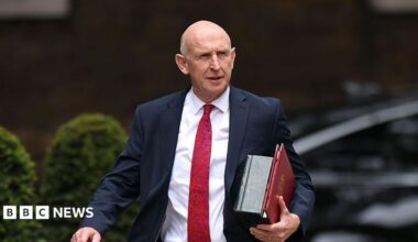 An image of John Healey from the waist up. He is walking and wearing a dark suit with a white shirt and red patterned tie. He is carrying a red folder. The background is blurred by you can see that he is outside from the outline of bushes.