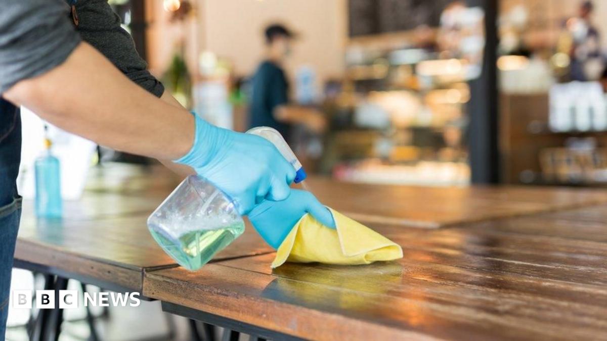Person wearing blue disposable gloves sprays cleaning solution onto a wooden table and wipes the surface with a yellow cloth inside a café or restaurant, with the background softly blurred.