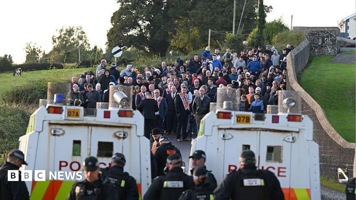 Two police Land Rovers parked in front of a crowd of people gathered on a country road.
