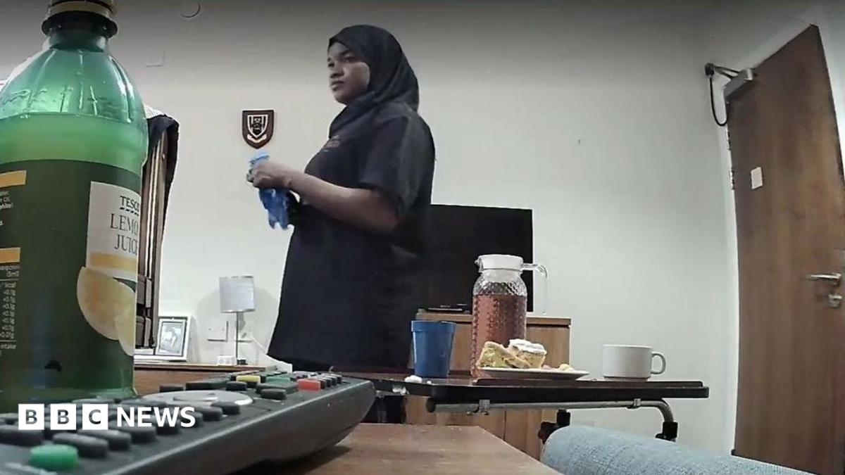 Sami is a young African woman in a black hijab in a clinical-looking room. Food can be seen on the table in front of her. The camera angle is low and looks to be set up secretly.