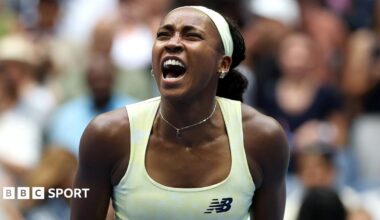 Coco Gauff, wearing a pale yellow vest, puts her left index finger behind her ear