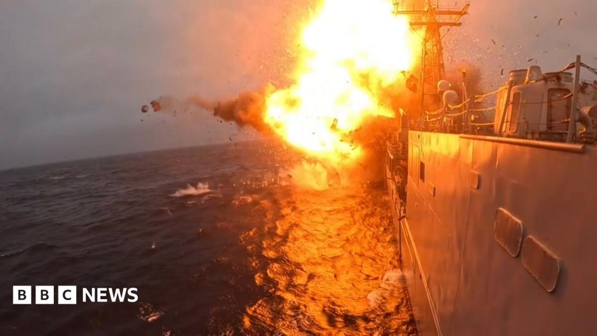 Orange flames and smokes erupts from an explosion on USS Klakring. Part of the grey warship is visible on the surface of a dark blue sea.
