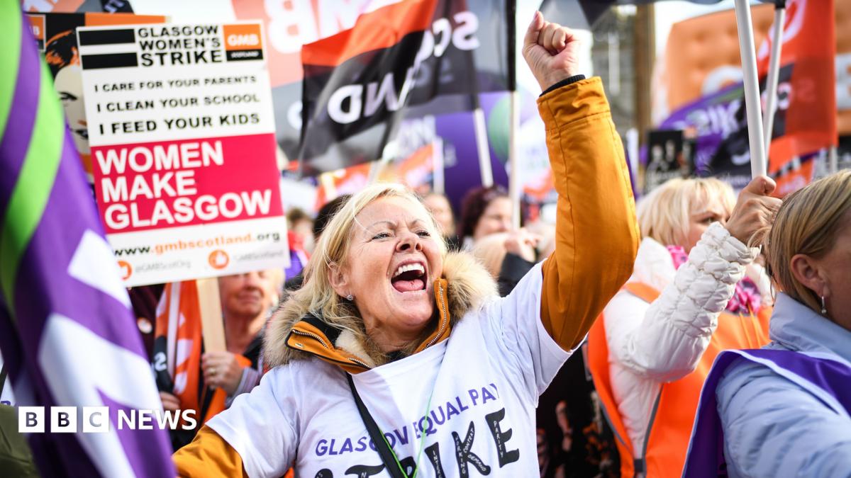 Protestors on a march. One woman is in the centre of the picture, punching the air and yelling. Many of the women around her are holding flags or placards, including one that says Glasgow Women's Strike and Women Make Glasgow.