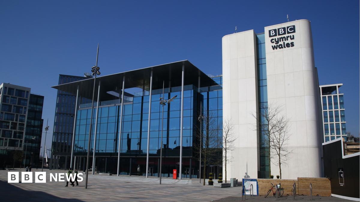 A huge glass building with pillars on central square. The white brick has black letters on it saying 'BBC Cymru Wales'. The background is a blue sky.