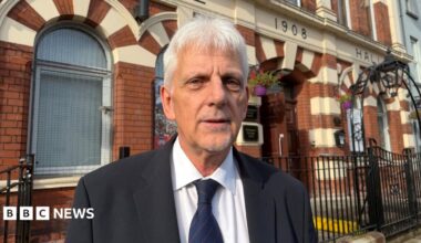 A white haired man looking at the camera. He is wearing a black jacket and white shirt. His tie is navy with red pin stripes.