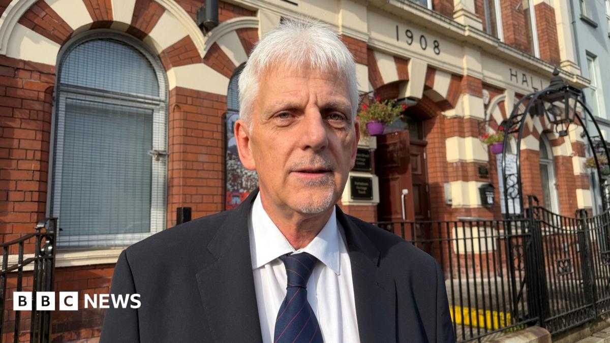 A white haired man looking at the camera. He is wearing a black jacket and white shirt. His tie is navy with red pin stripes.