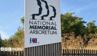 A large white sign that has the words "National Memorial Arboretum" on it with in smaller lettering "Part of Royal British Legion'. In the background is greenery and a fence.